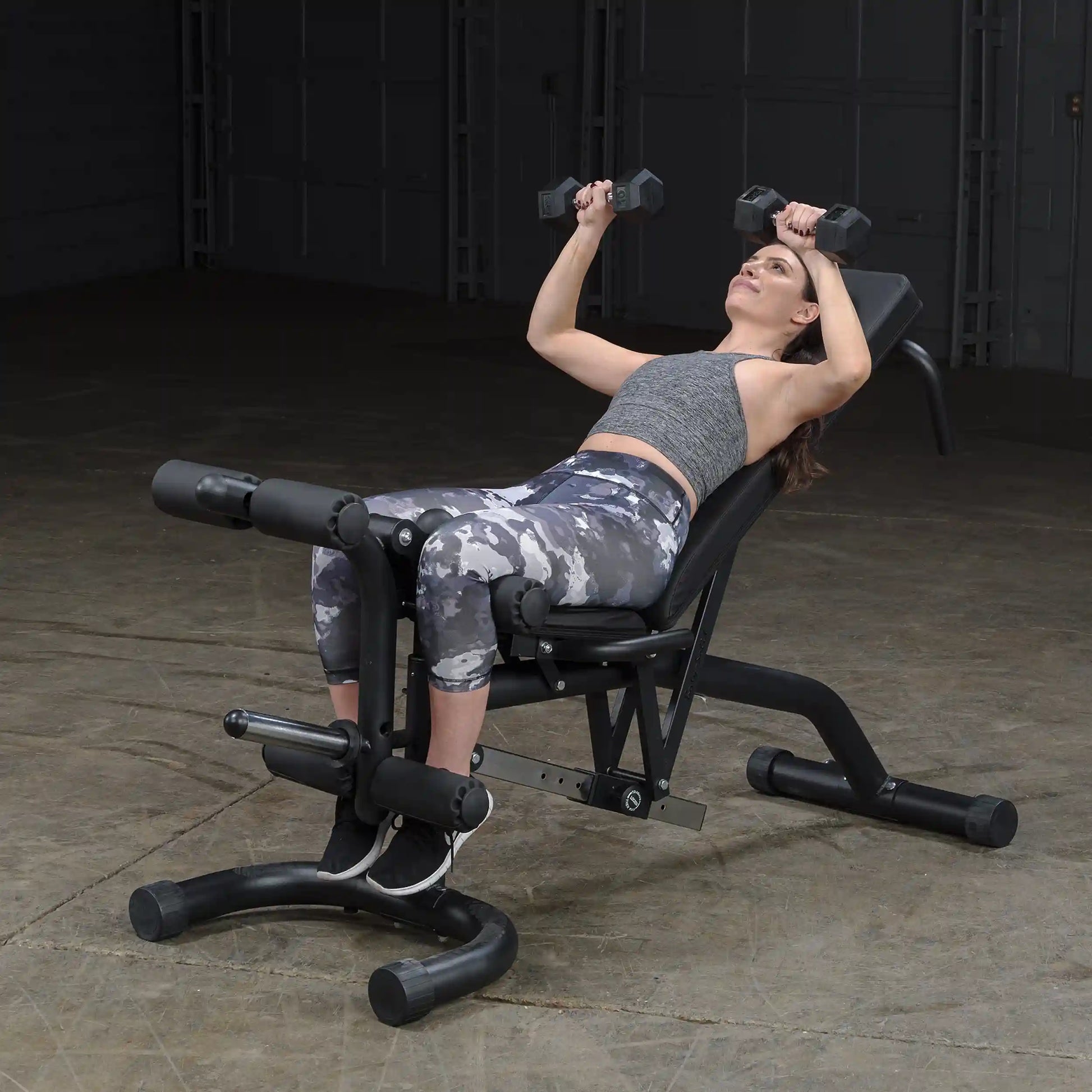 Woman using weight bench for dumbbell press in gray camo leggings.