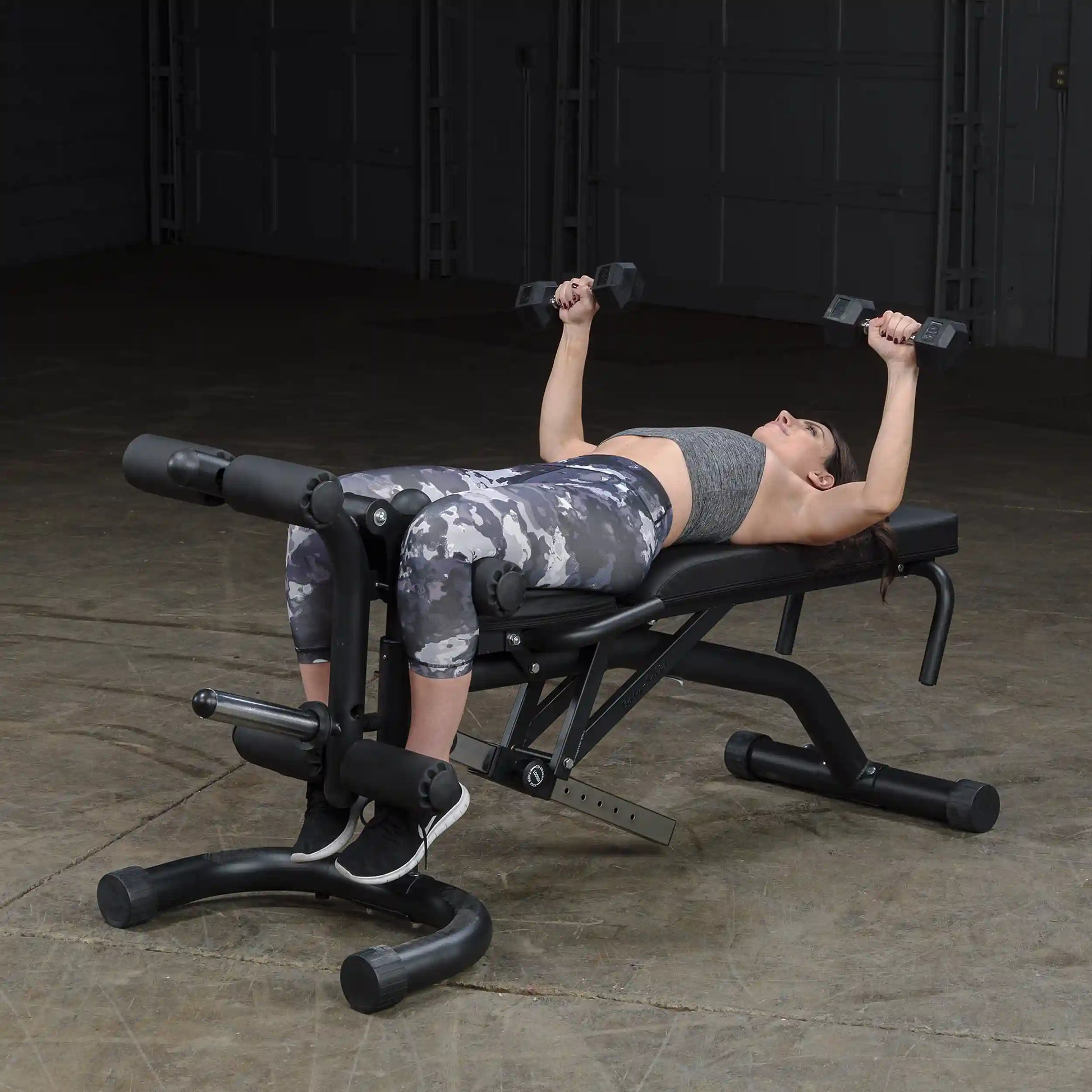 Woman using a weight bench with dumbbells, wearing gray camo leggings, in a gym setting.