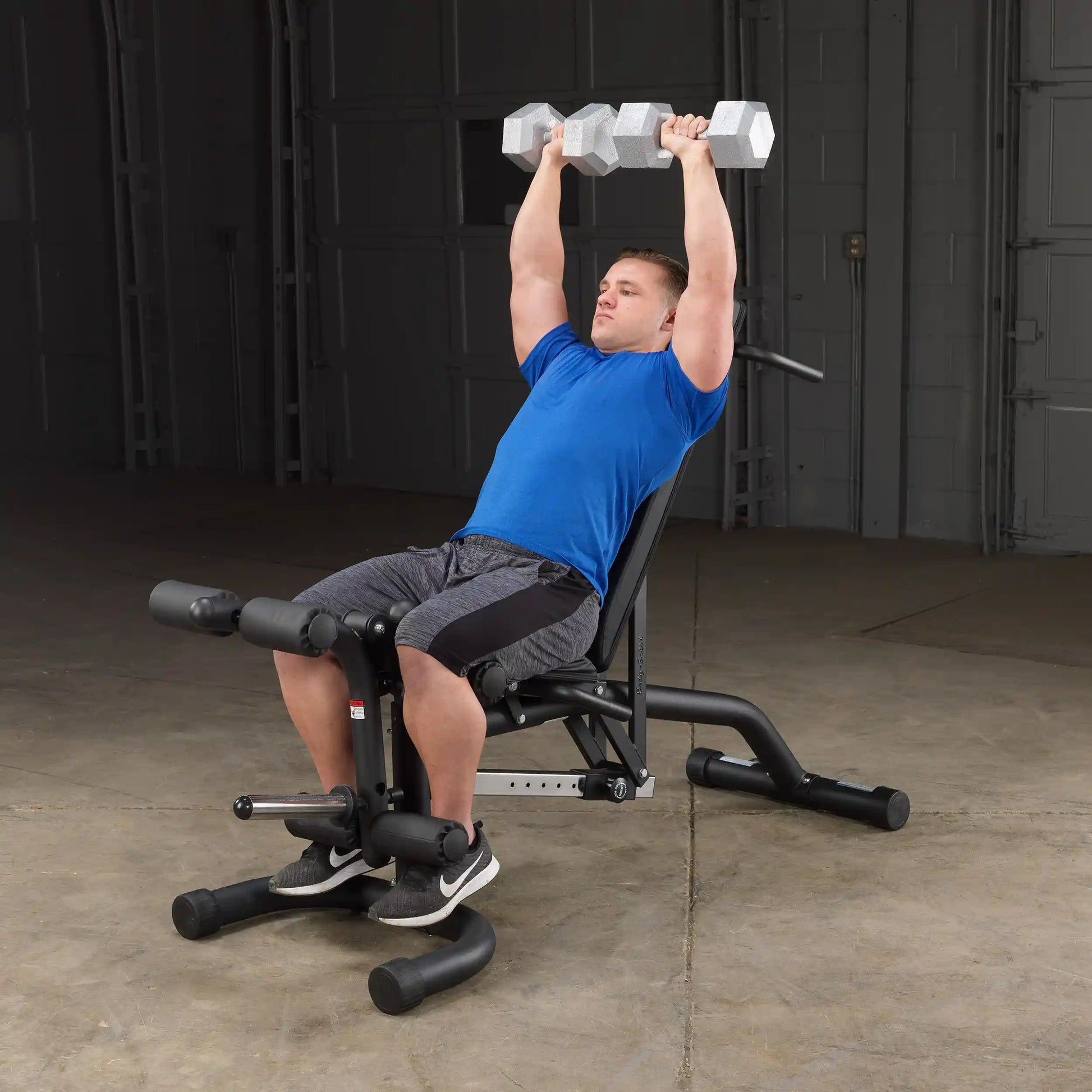 Man in blue shirt lifting dumbbells on adjustable workout bench in gym.