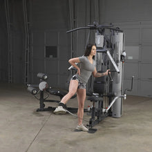 Woman exercising on Body-Solid home gym, performing leg curl with ankle strap.