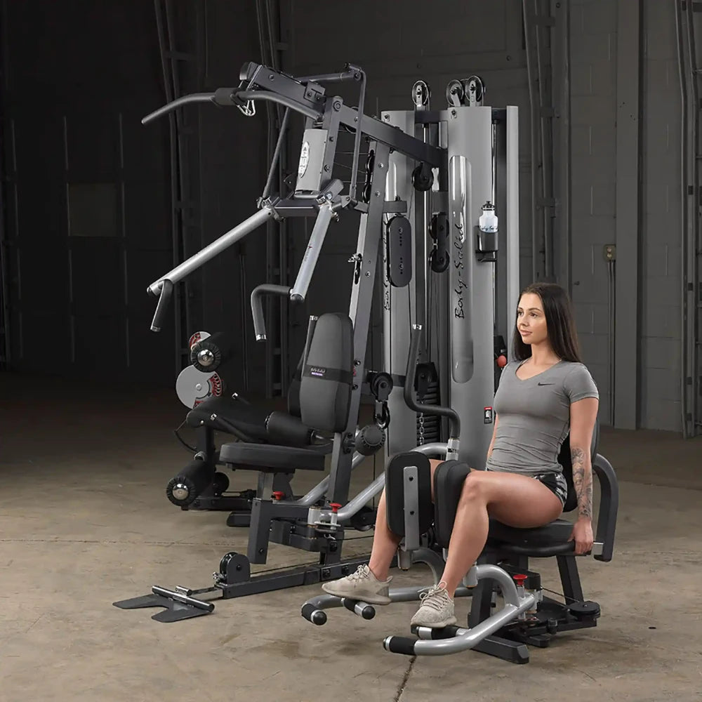 Woman using Body-Solid home gym machine for leg exercises in a gray workout outfit.