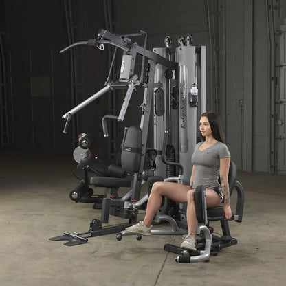 Woman using Body-Solid home gym machine for leg exercises in a gray sports outfit.
