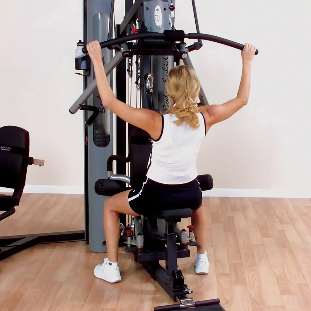 Woman using Body-Solid home gym for lat pulldown exercise on a wooden floor.