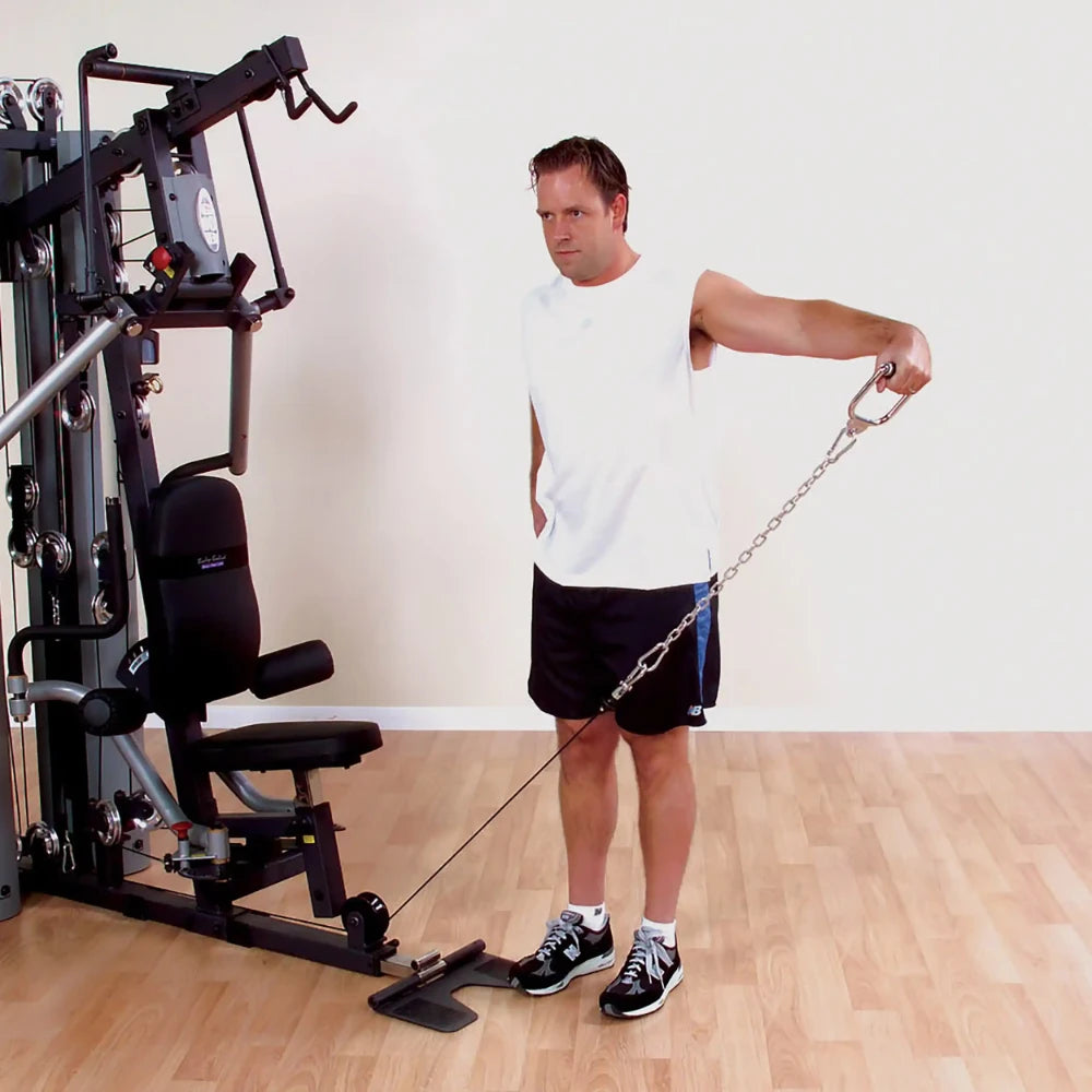 Man using a cable machine for lateral raises in a gym, wearing a white tank top and black shorts.