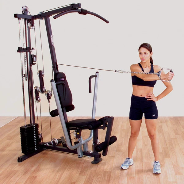 Woman using a black home gym machine for cable exercises on a wooden floor.