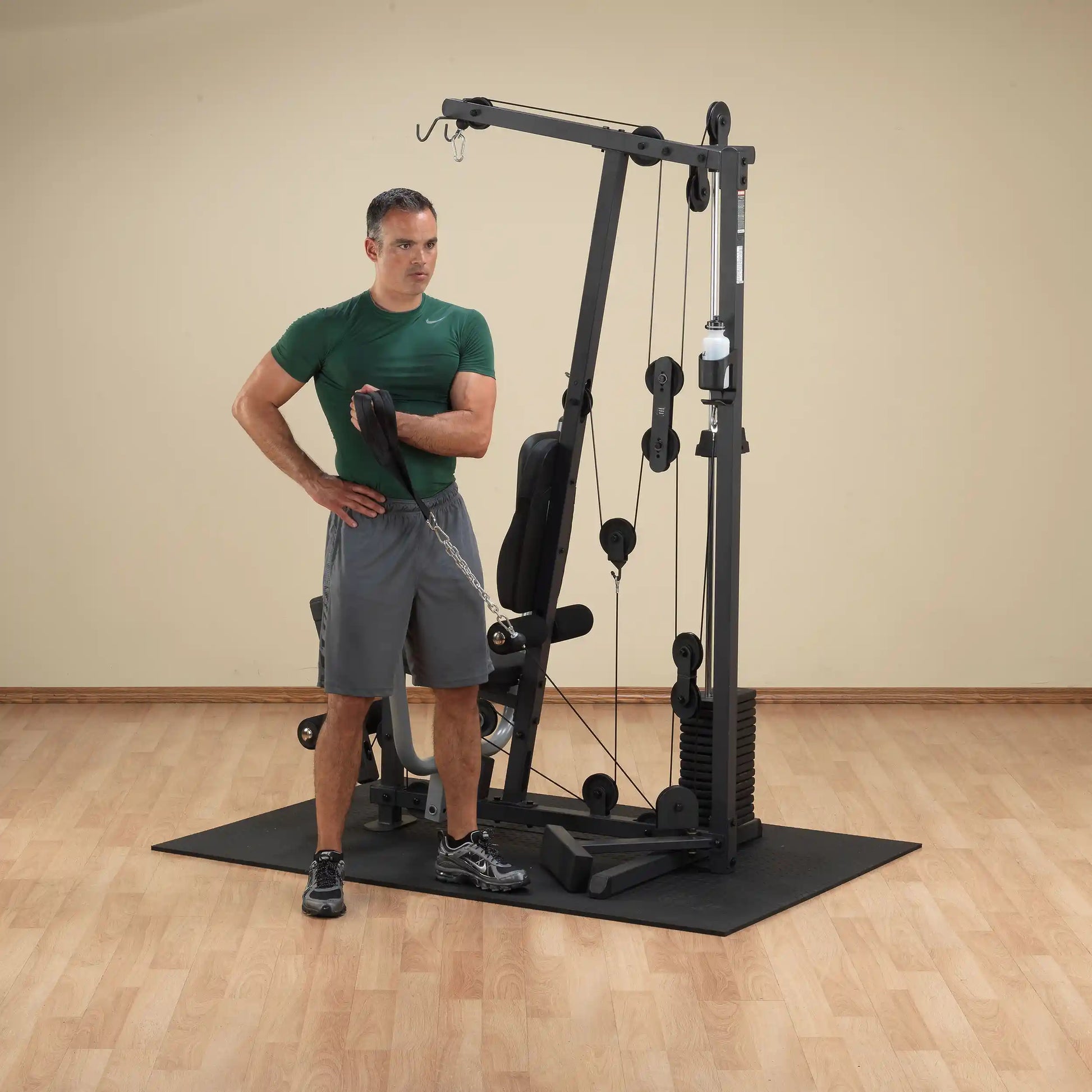 Man using a home gym machine with pulley system for arm workout, wearing green shirt and gray shorts.