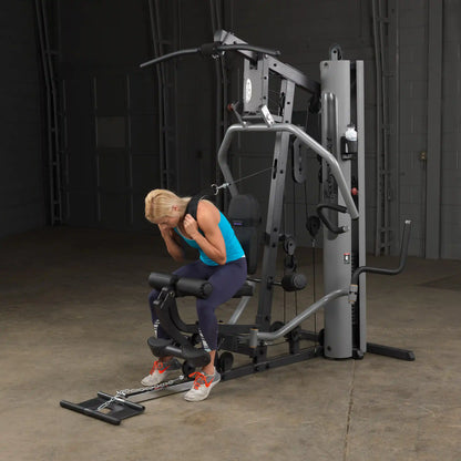 Woman exercising on Body-Solid home gym machine with lat pulldown bar in a dimly lit room.