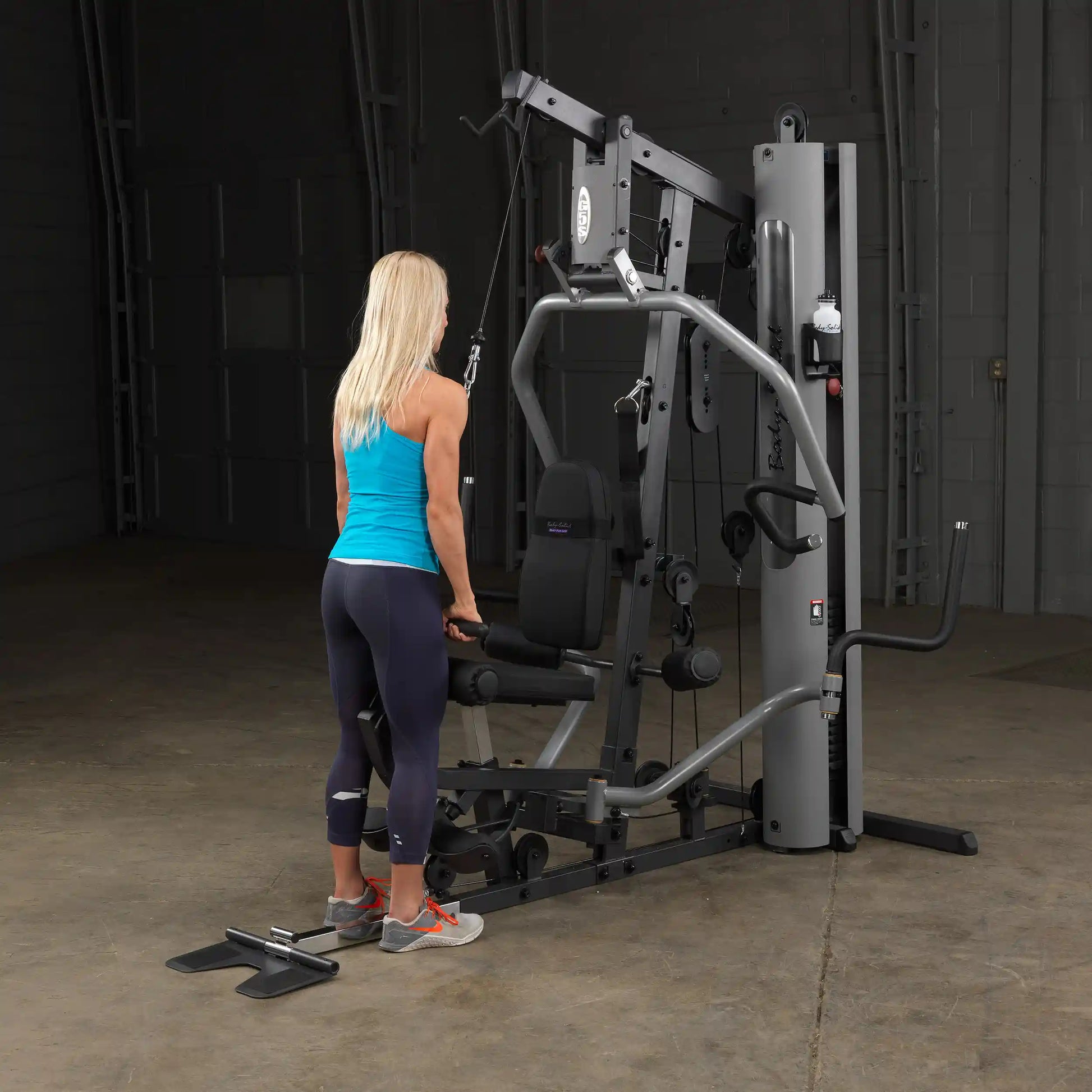 Woman using Body-Solid home gym in bright blue tank, performing cable exercise.