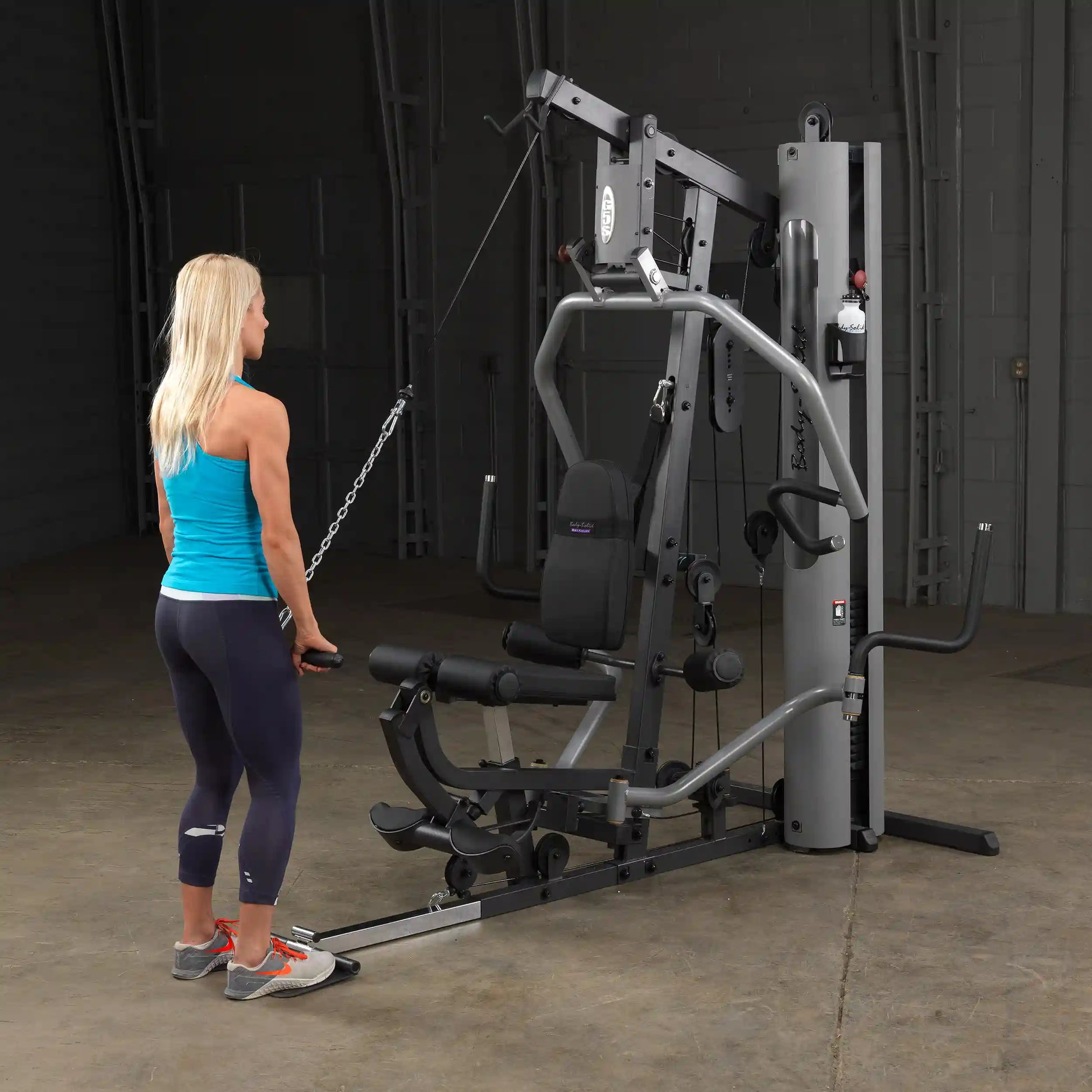 Woman using Body-Solid home gym in a blue tank top, performing cable exercises for strength training.