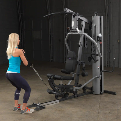 Woman using Body-Solid home gym in blue tank top, performing cable exercises.