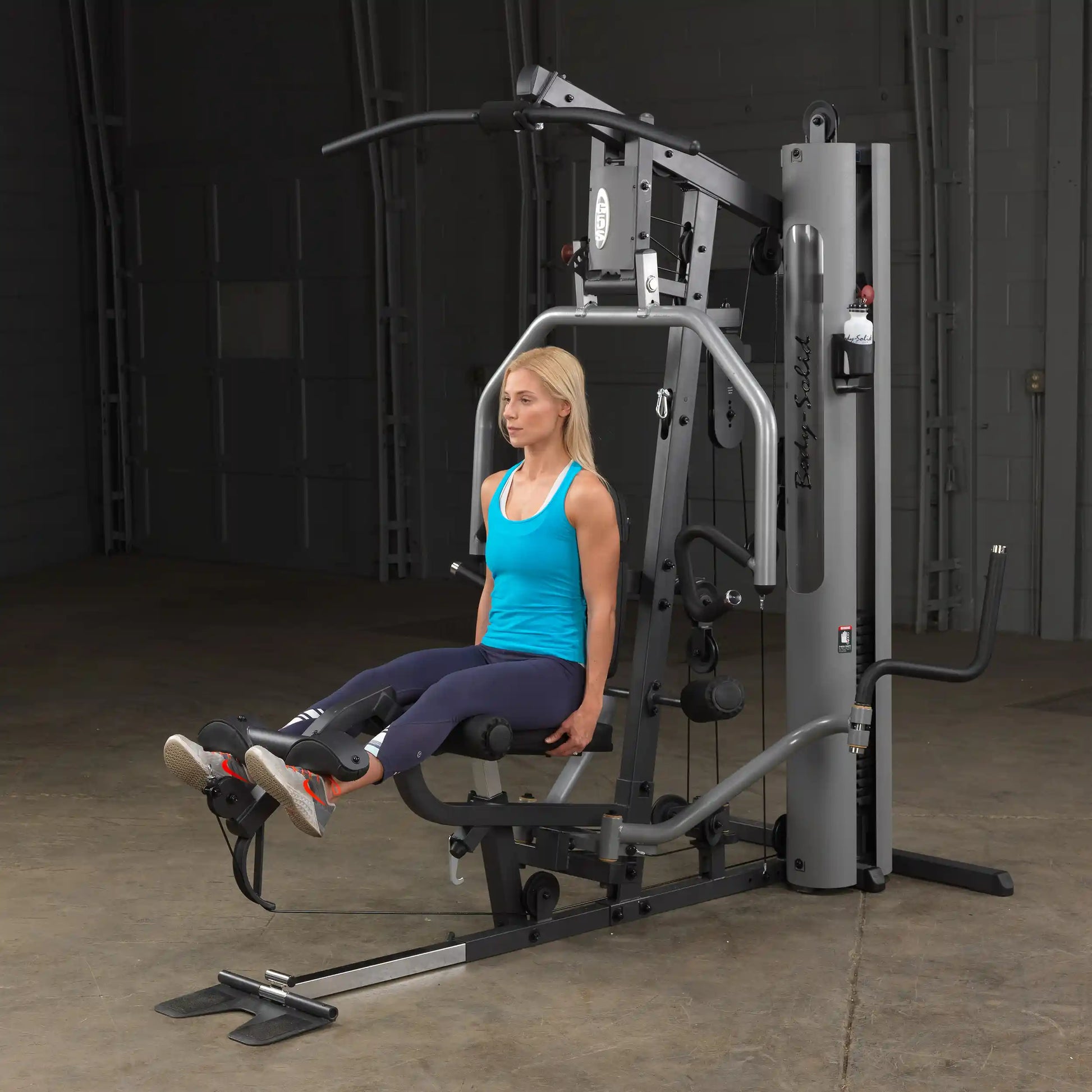Woman using Body-Solid gym machine in blue tank top, performing leg exercises.