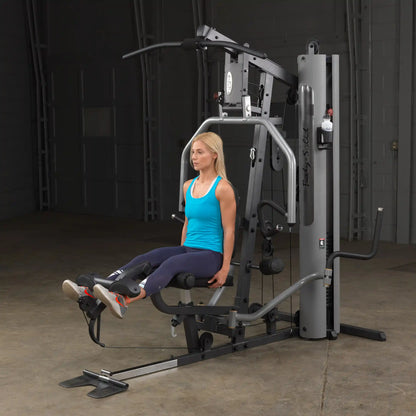 Woman using Body-Solid gym machine in blue tank top, performing leg exercises.