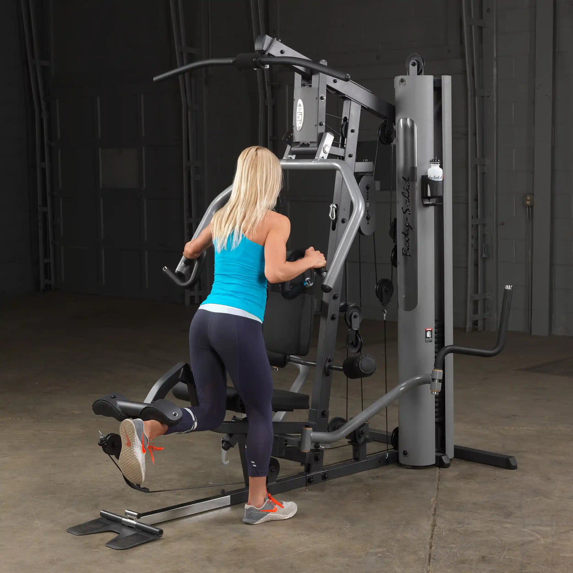 Woman using Body-Solid home gym in blue tank top for leg workout.