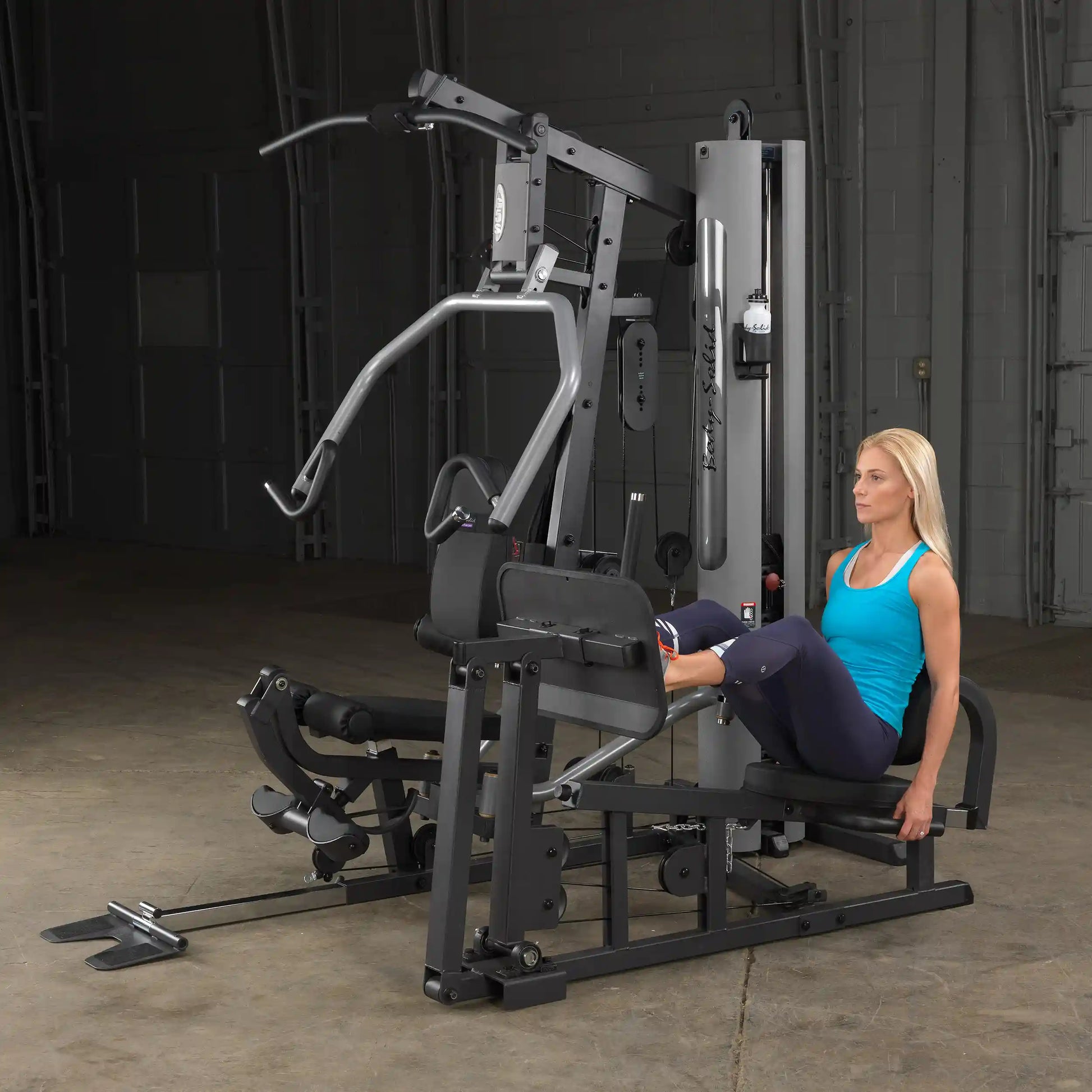Woman using Body-Solid G6BR home gym, performing leg press in a blue tank top.