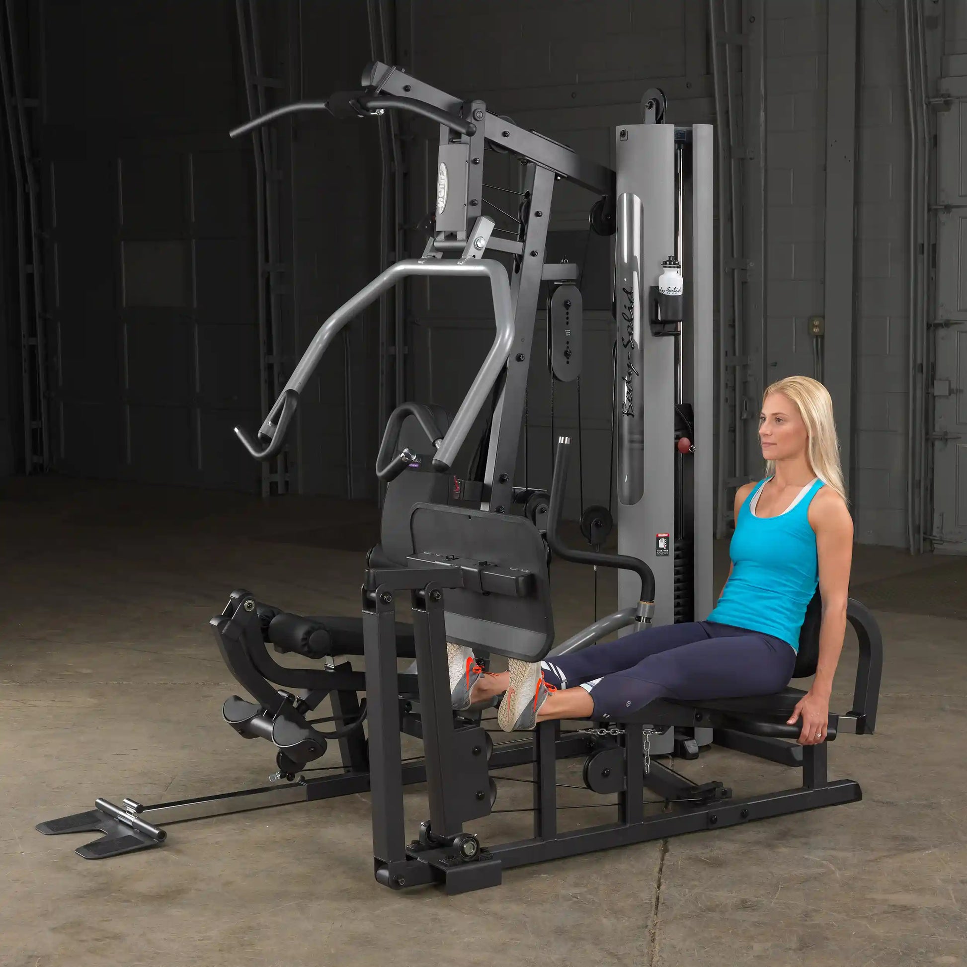 Woman using a Body-Solid home gym in blue tank top performing leg press exercise.