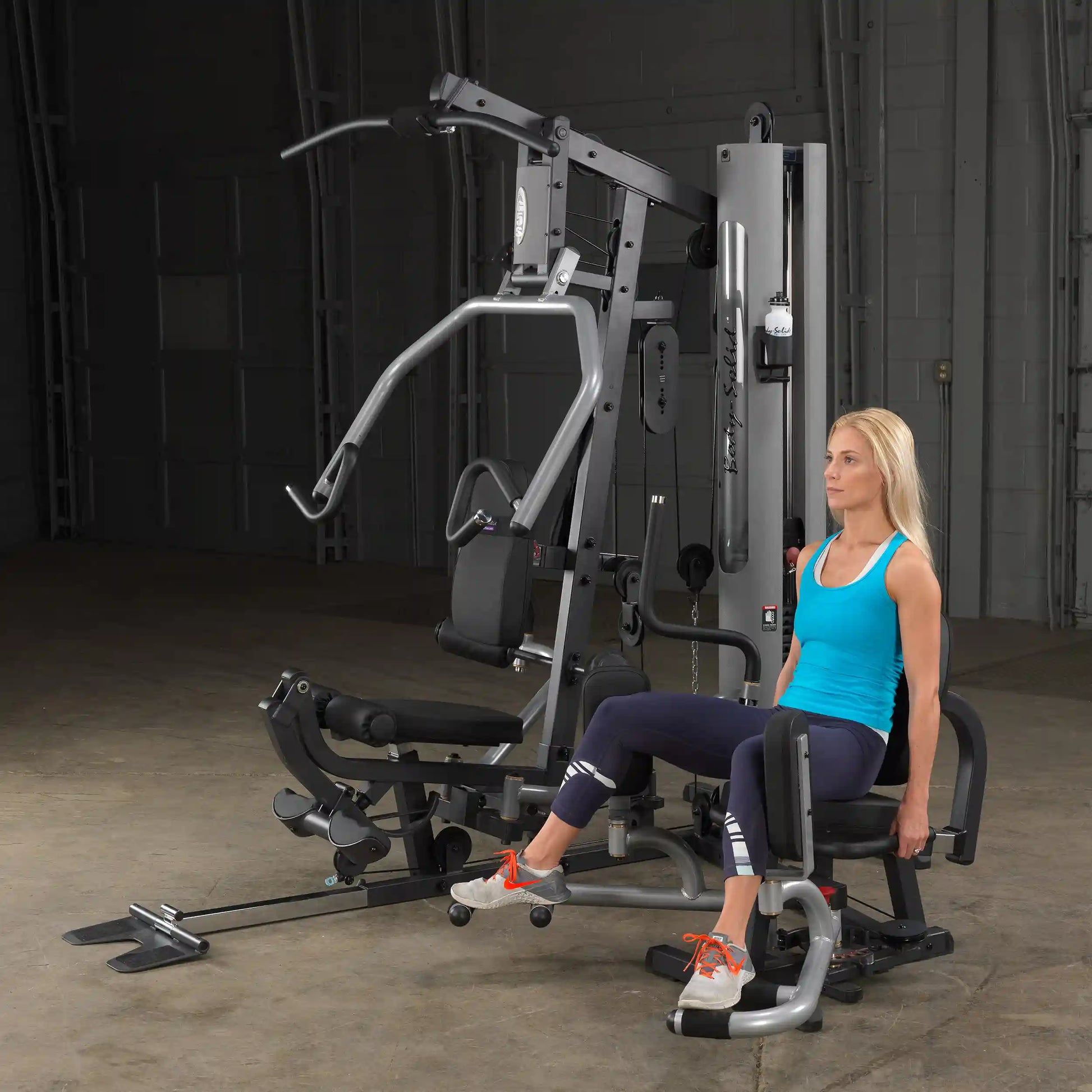 Woman using Body-Solid home gym equipment in a blue tank top, performing leg exercises.