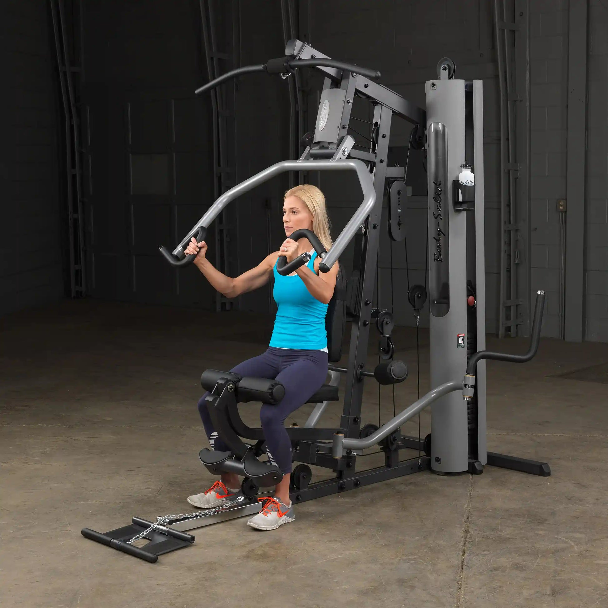 Woman using Body-Solid home gym in blue tank top, performing chest press exercise.
