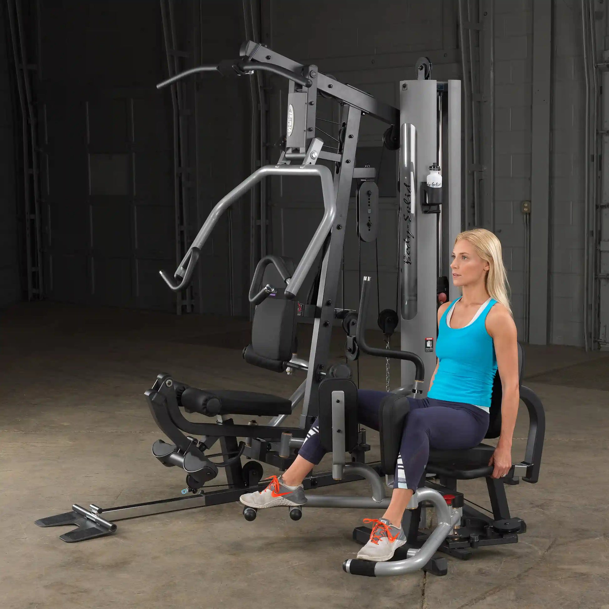 Woman using Body-Solid gym equipment in blue tank top for leg exercise.