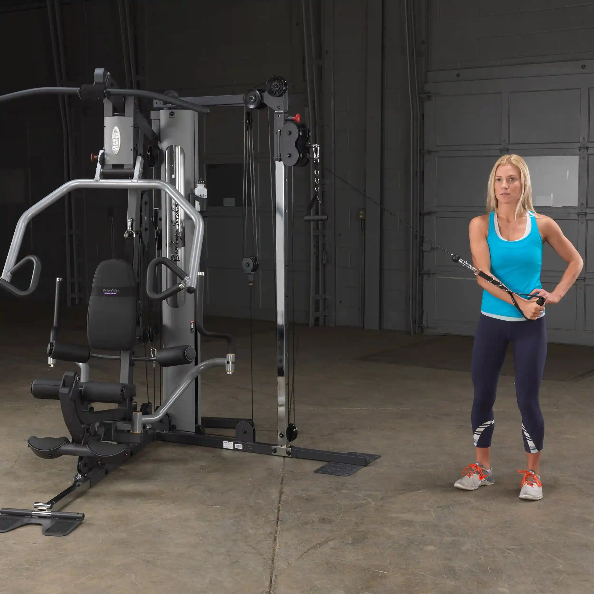 Woman using a Body-Solid home gym machine for cable exercises in a blue tank top.