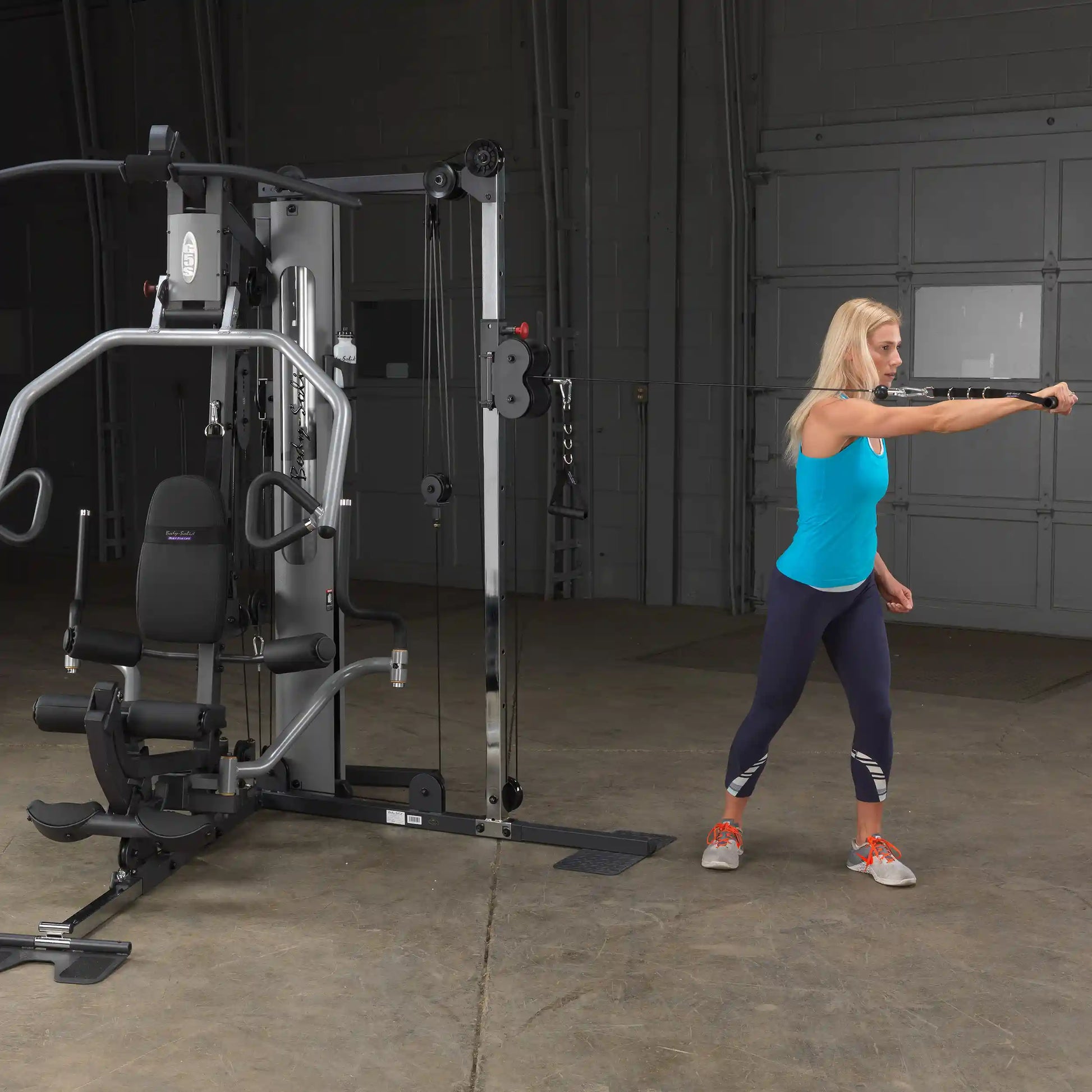 Woman using a Body-Solid home gym in a blue tank top, performing a cable exercise.