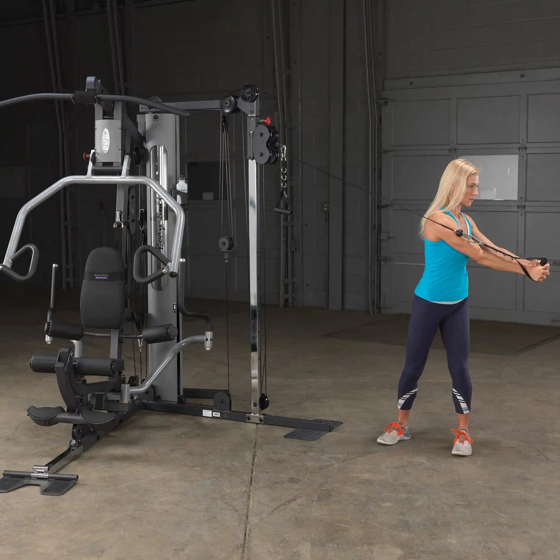 Woman using Body-Solid home gym machine with cable pulleys in a garage setting.