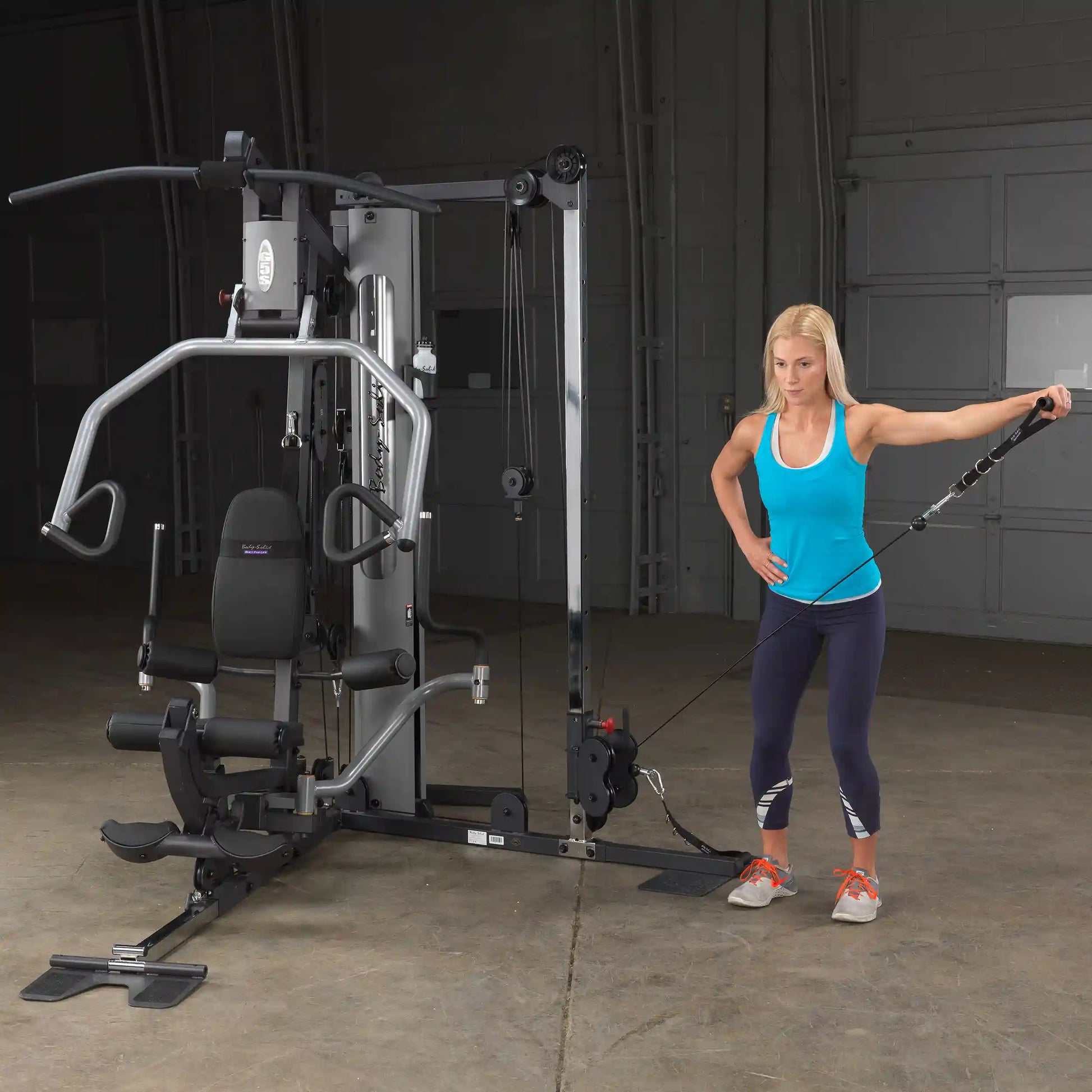 Woman using Body-Solid gym machine for cable exercises in a blue tank top.