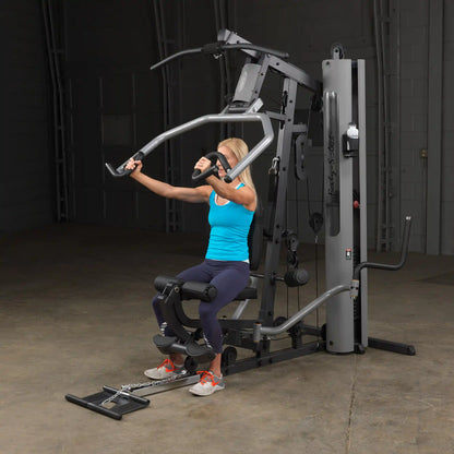 Woman using Body-Solid home gym in blue tank, performing chest press.