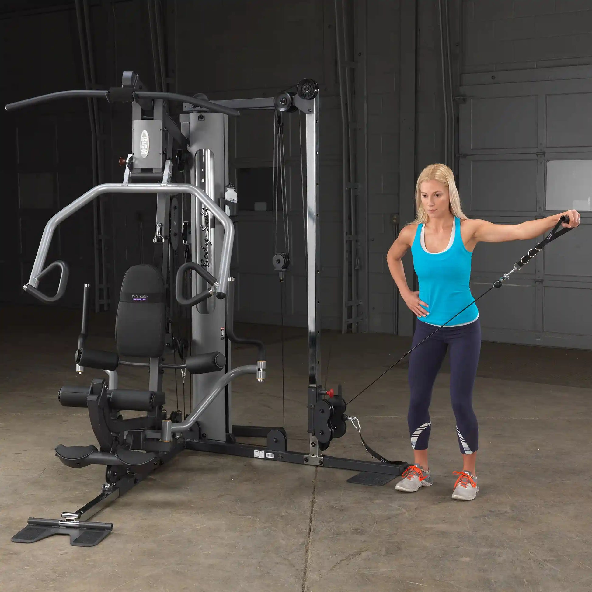 Woman using Body-Solid home gym in blue tank top, demonstrating cable exercise.