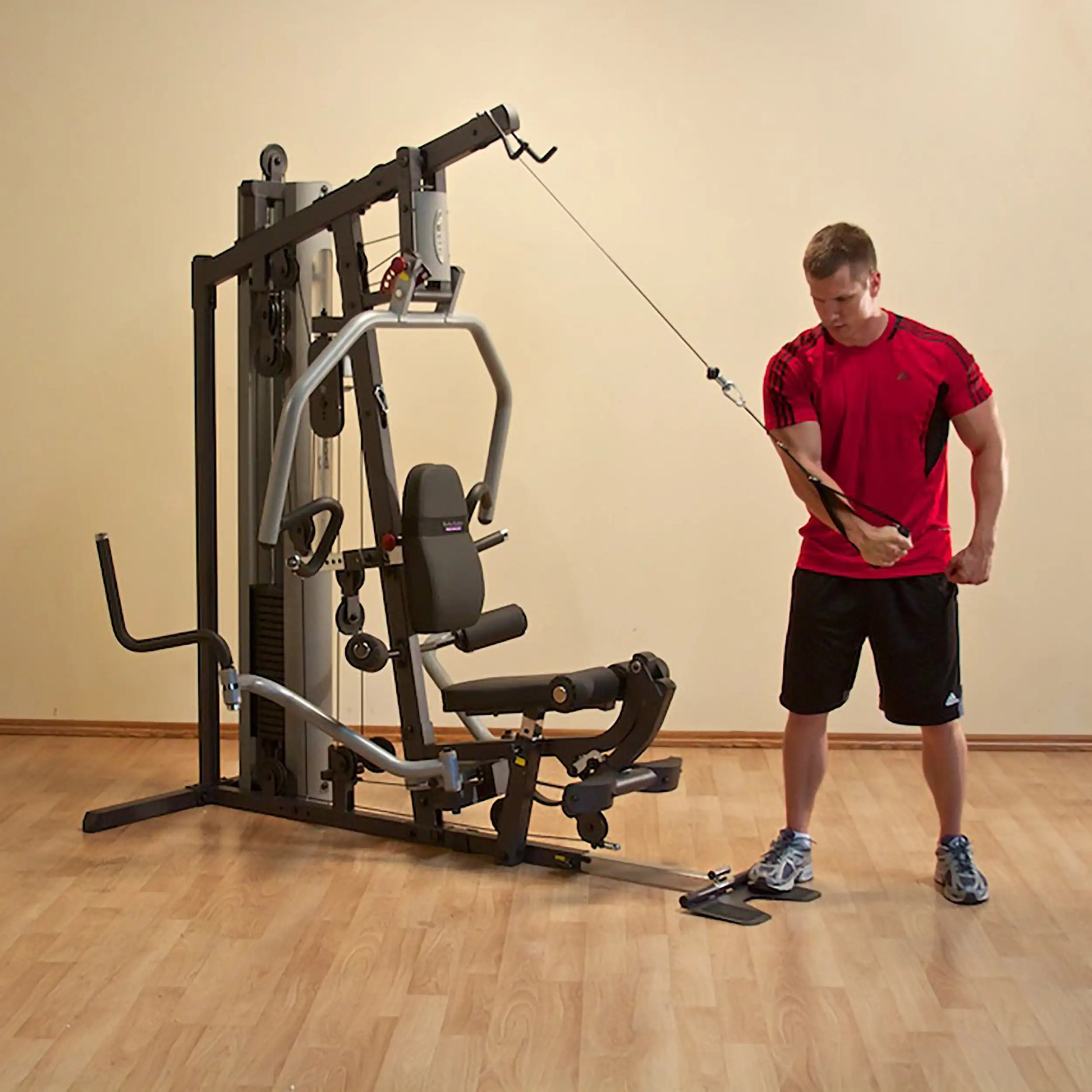 Man using a home gym machine for cable exercises, wearing a red shirt and black shorts.
