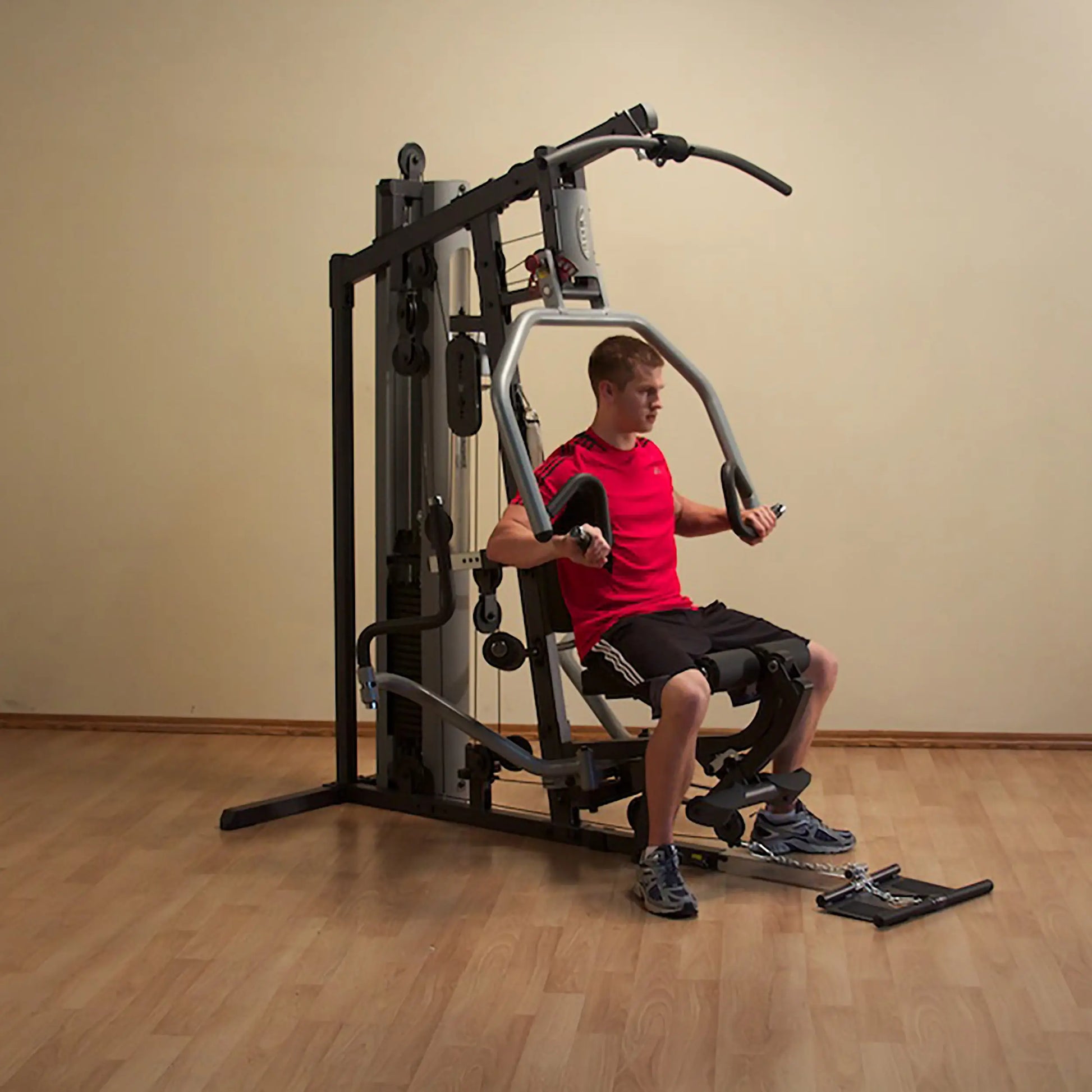 Man using home gym machine in red shirt, performing chest press on wooden floor.