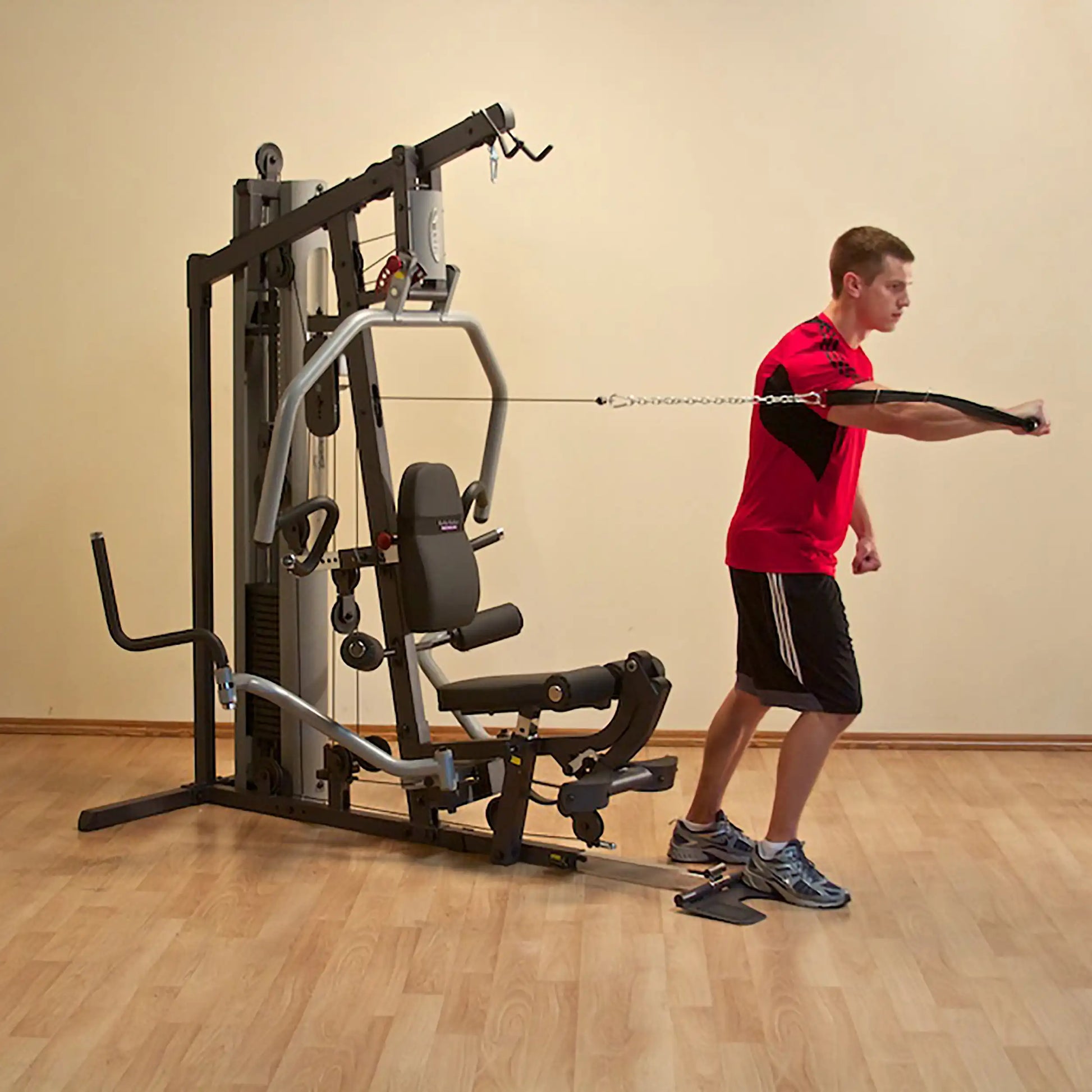 Man exercising with a home gym machine, wearing a red shirt and black shorts on a wooden floor.