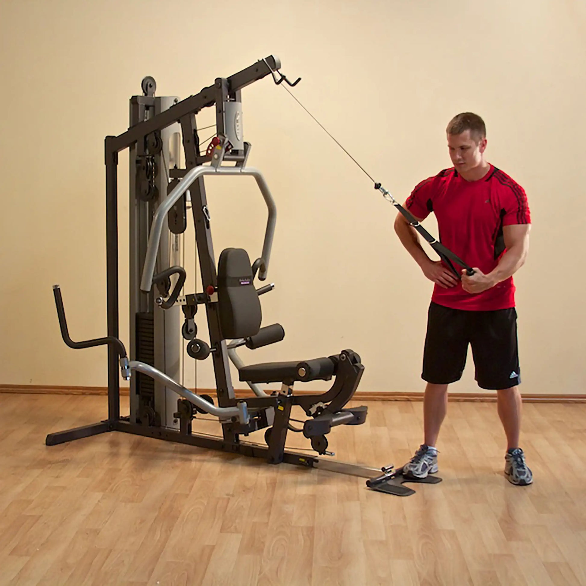 Man in red shirt using a home gym machine with cable attachment on wooden floor.