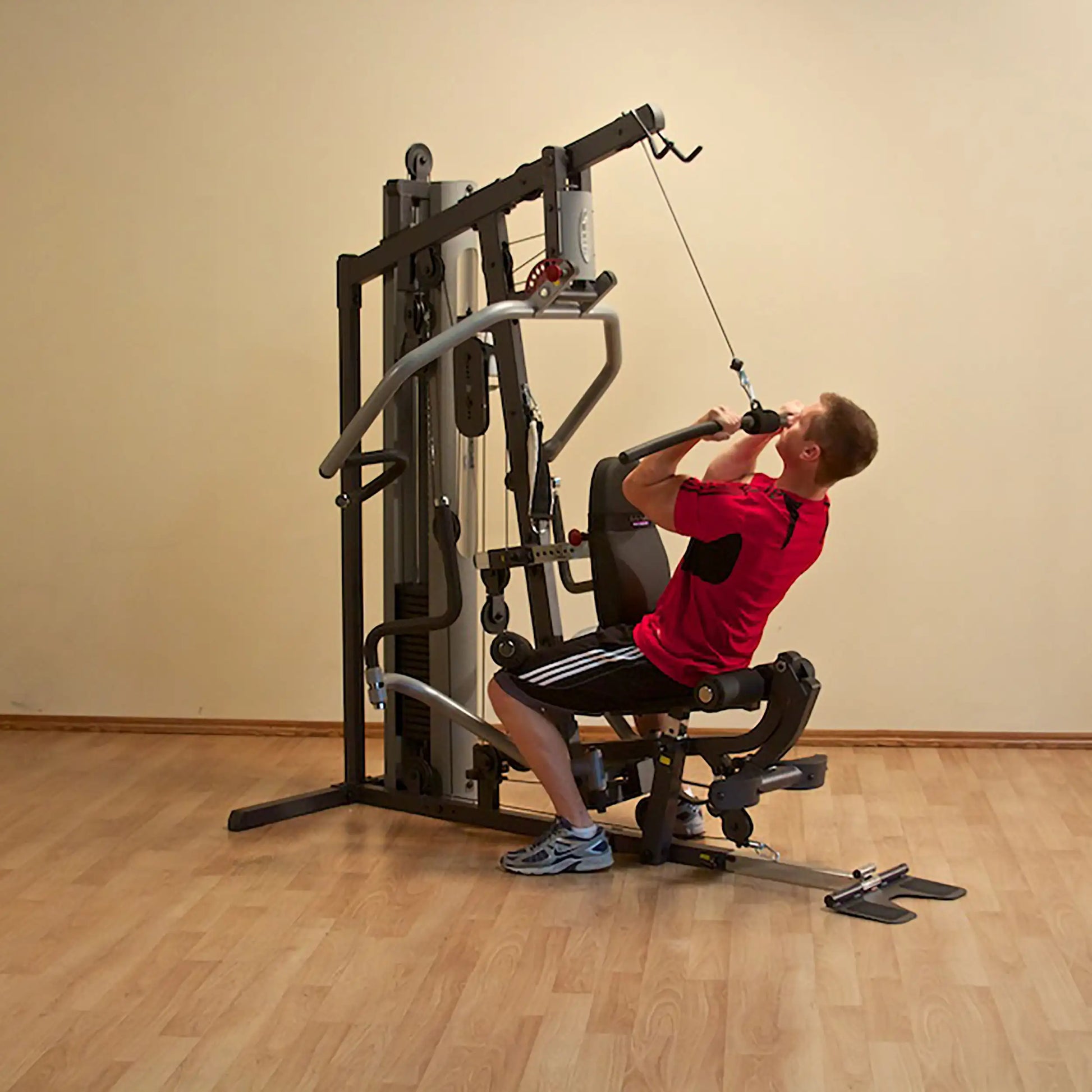 Man using a home gym machine with a lat pulldown attachment, wearing red shirt and black shorts.