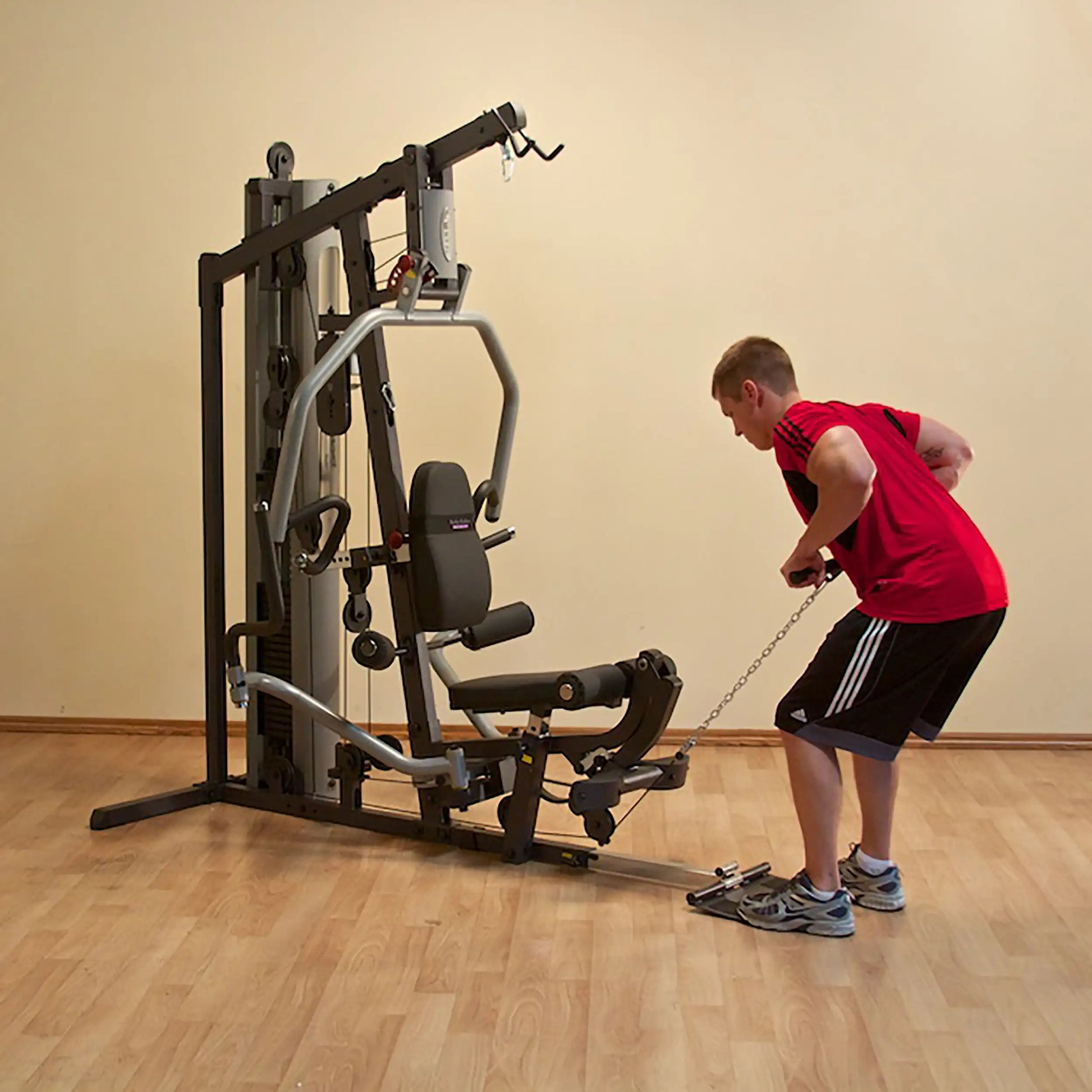 Man using a home gym machine in red shirt, performing a cable row exercise on wooden floor.
