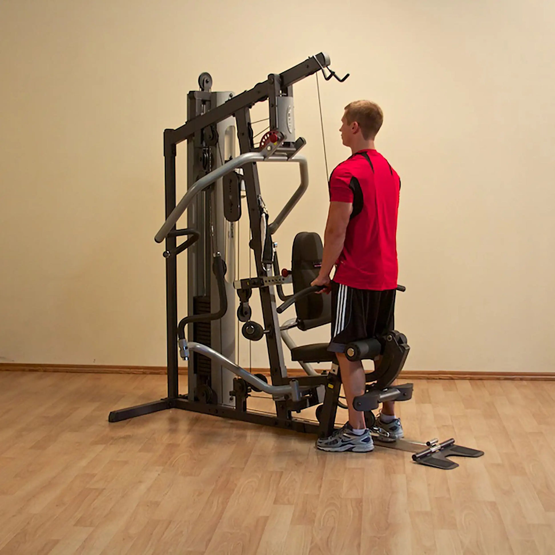 Man using multifunctional gym machine in red shirt and black shorts on wooden floor.
