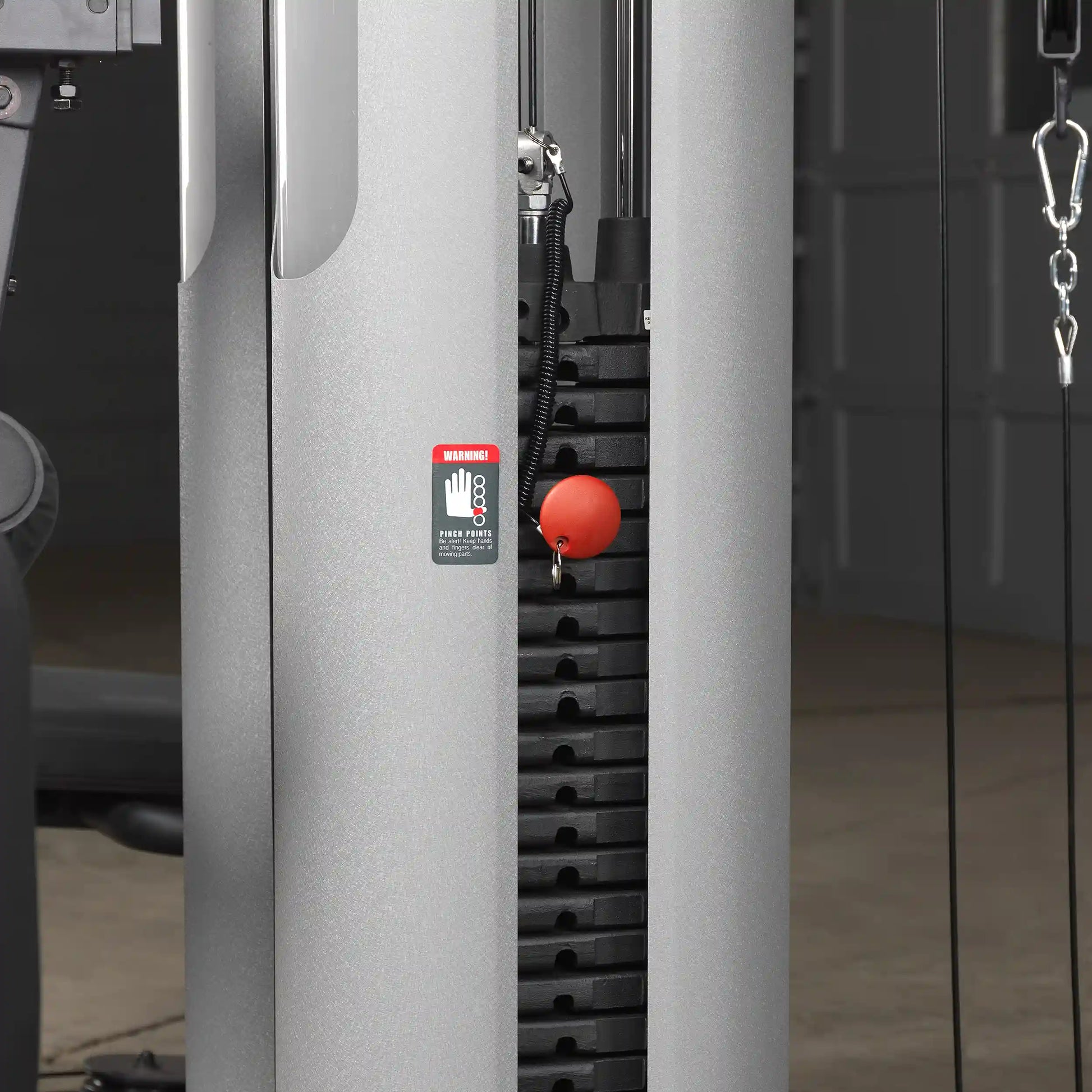 Weight stack of a cable machine with red pin and warning label in a gym setting.