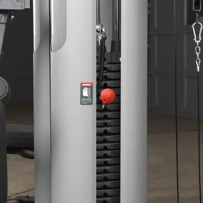 Weight stack of a cable machine with red pin and warning label in a gym setting.