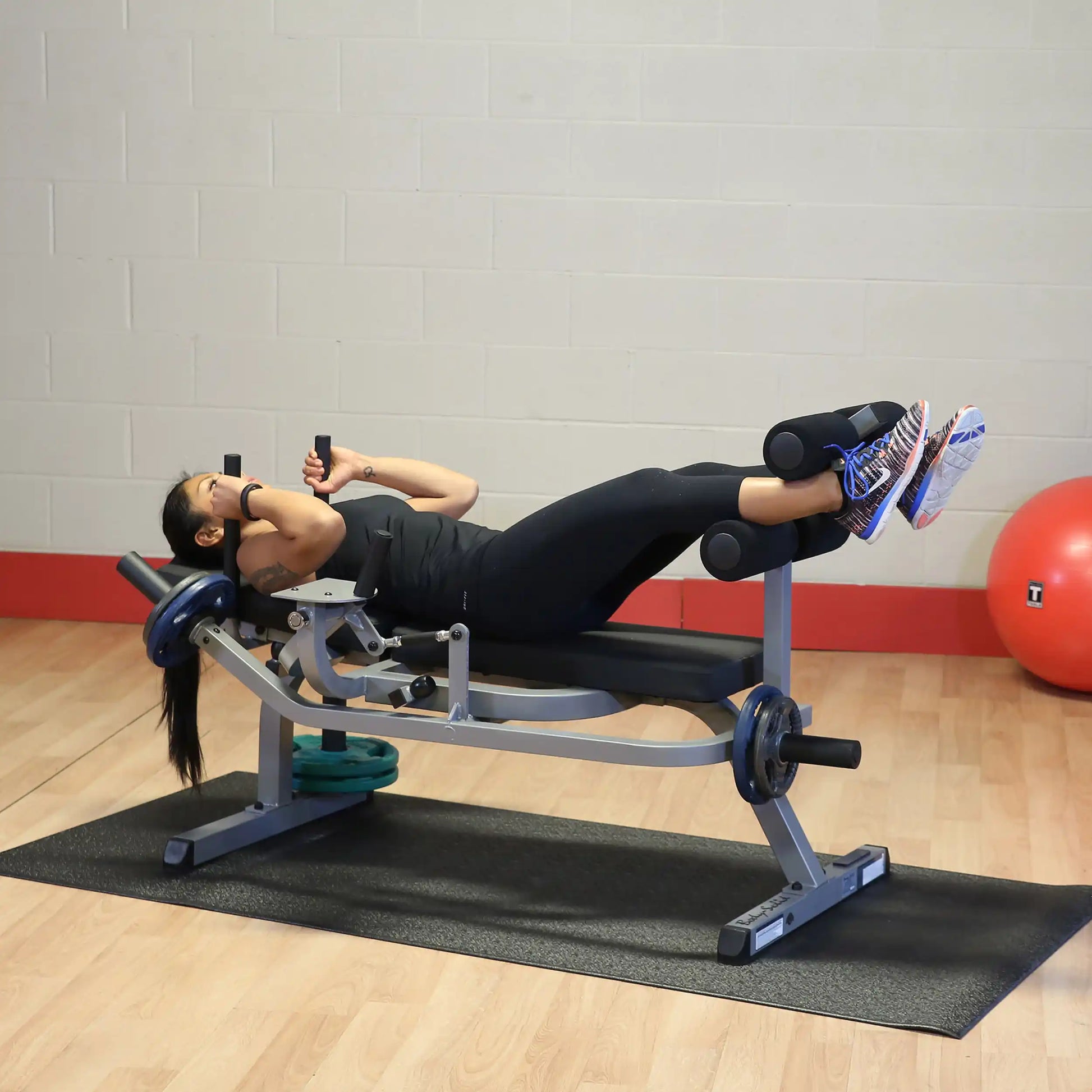 Woman using a Valor Fitness leg curl machine in a gym with red walls and wooden floor.