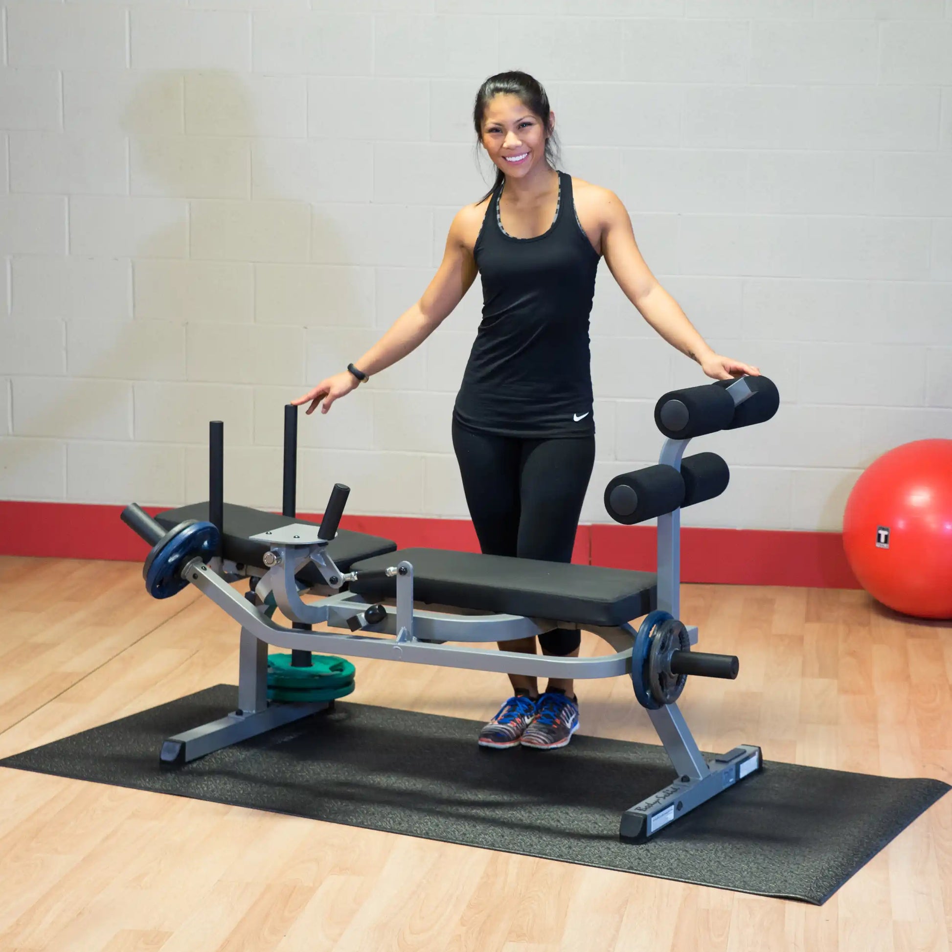 Woman in black workout attire standing by a gym leg extension machine with weights, smiling.