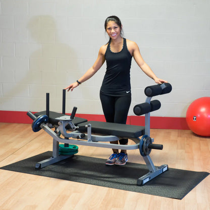Woman in black workout attire standing by a gym leg extension machine with weights, smiling.