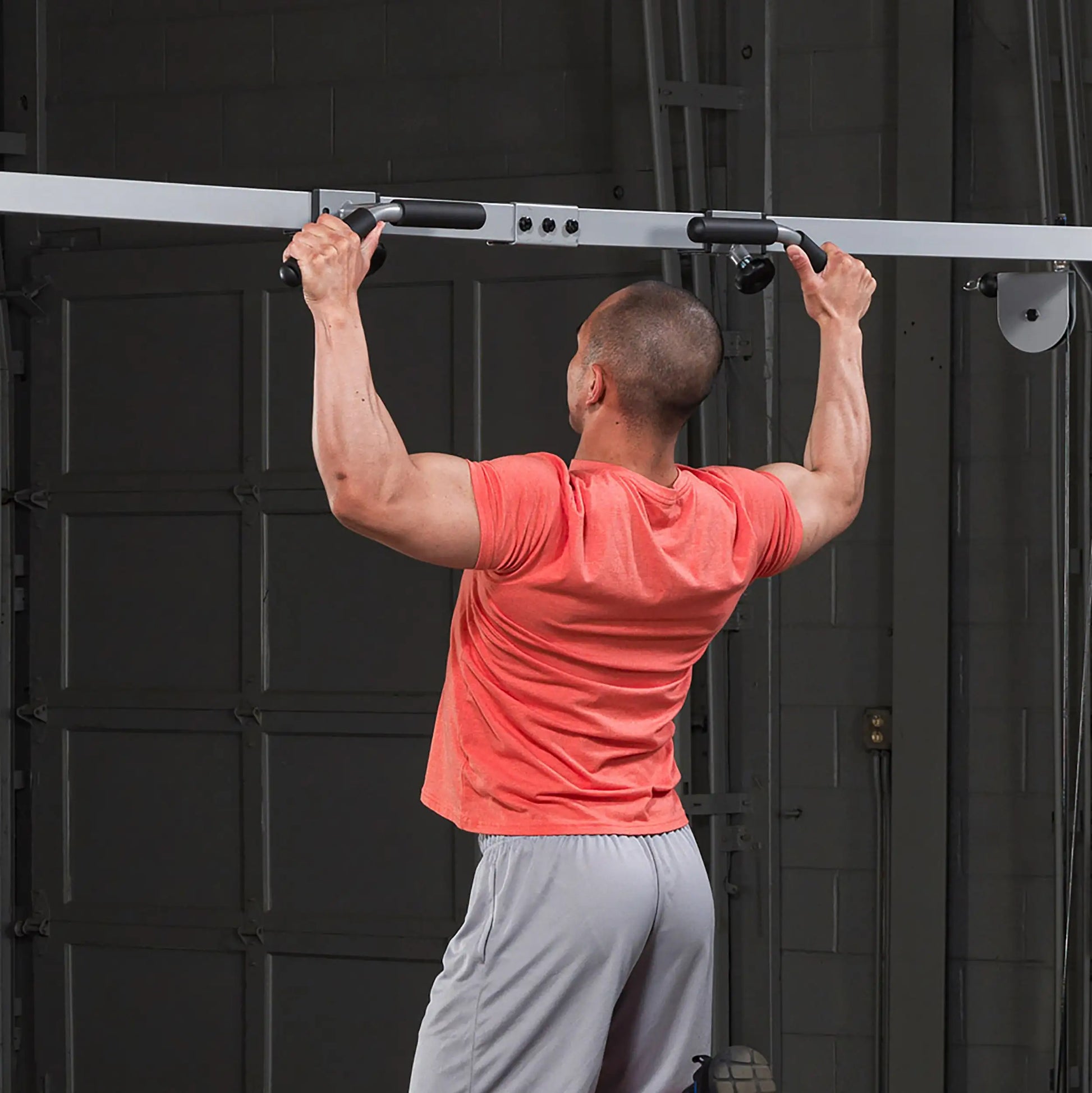 Man in red shirt doing pull-ups on a black pull-up bar in a garage gym.