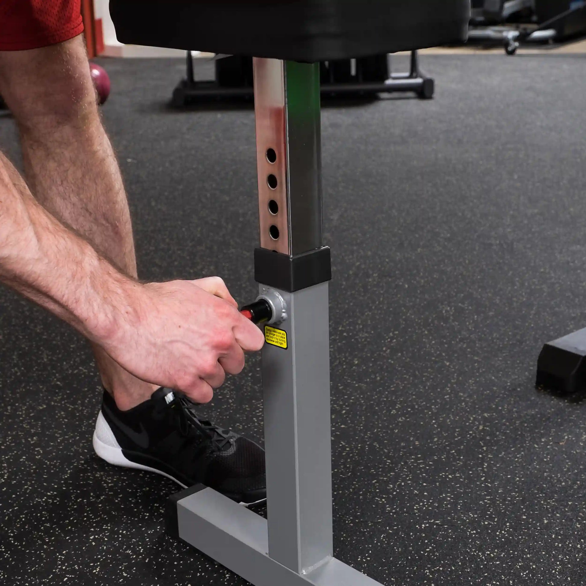 Adjustable gym bench leg being secured by a person in black sneakers on a rubber gym floor.