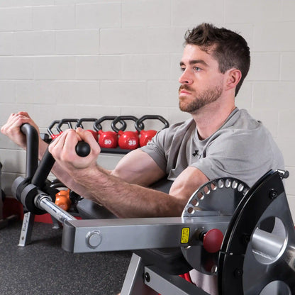 Man using a Rogue Fitness arm curl machine, focusing on bicep workout with red kettlebells in the background.