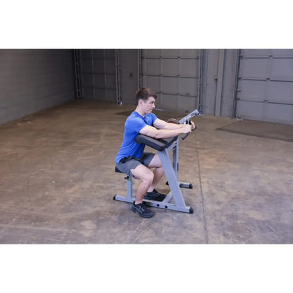Man using a seated preacher curl machine in a gym, wearing a blue shirt and gray shorts.
