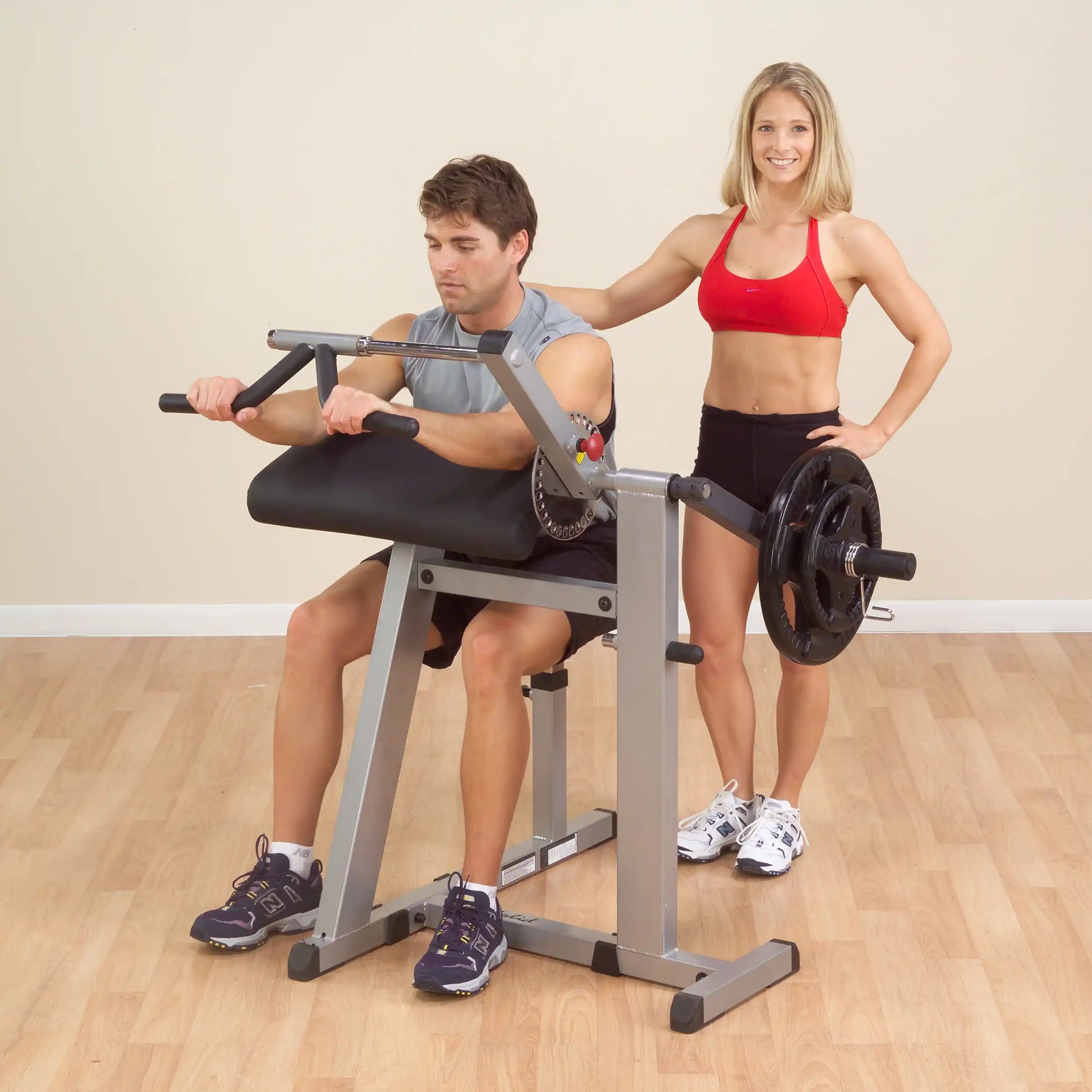 Man using Preacher Curl Bench with weights, assisted by woman in red sports bra.