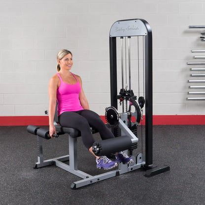 Woman using Body-Solid leg extension machine in gym, wearing pink top and black leggings.