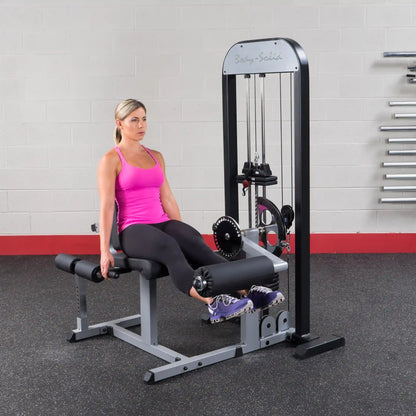 Woman using Body-Solid leg extension machine in gym, wearing pink top and black leggings.