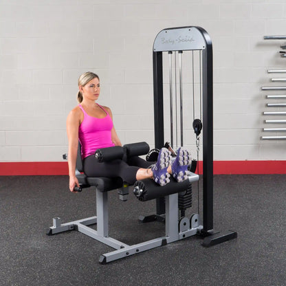 Woman using Body-Solid leg extension machine in gym, wearing pink top and black leggings.
