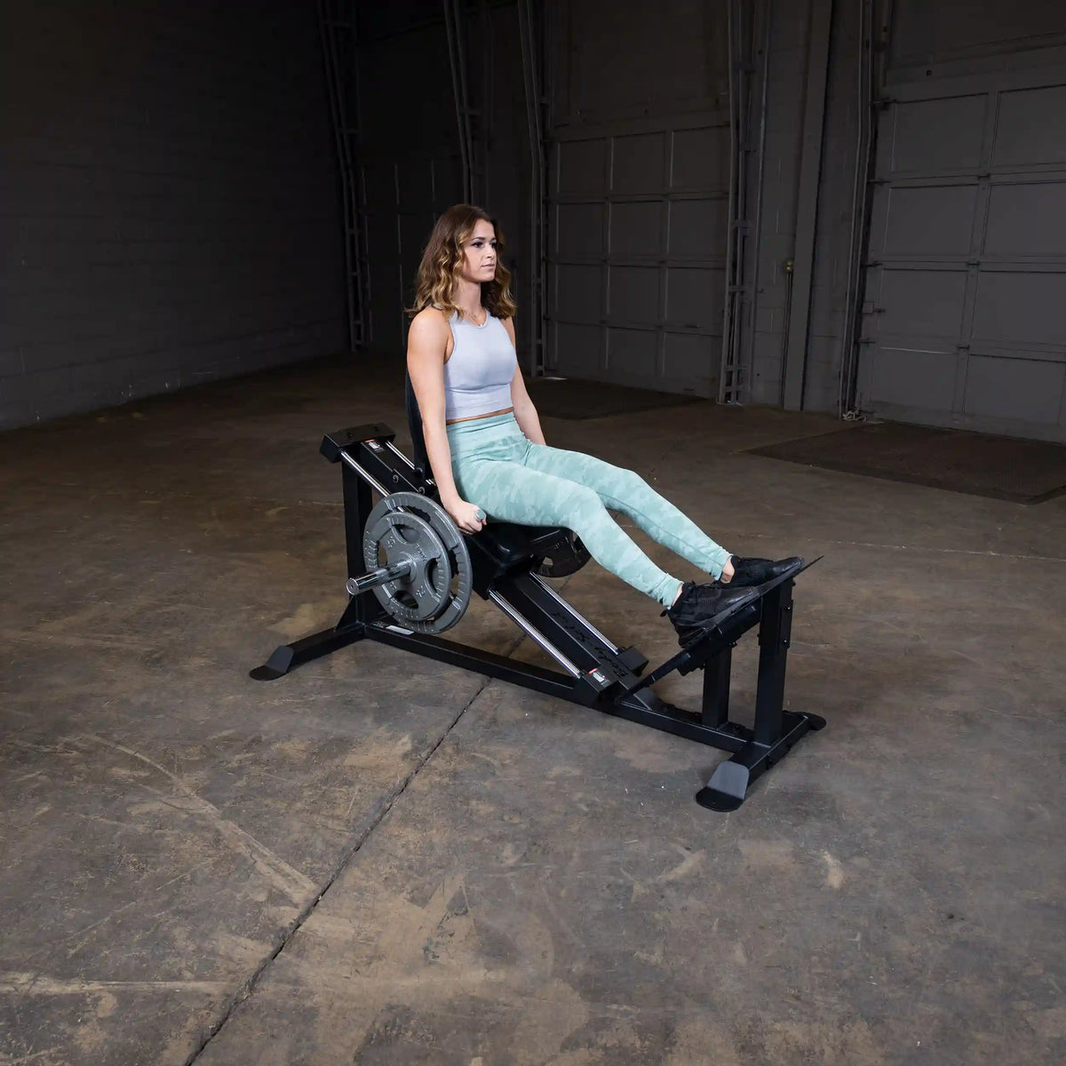 Woman using a leg press machine in a gym, wearing light green leggings and a gray tank top.