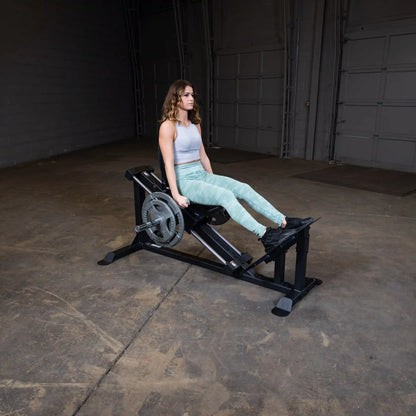 Woman using a leg press machine in a gym, wearing light green leggings and a gray tank top.